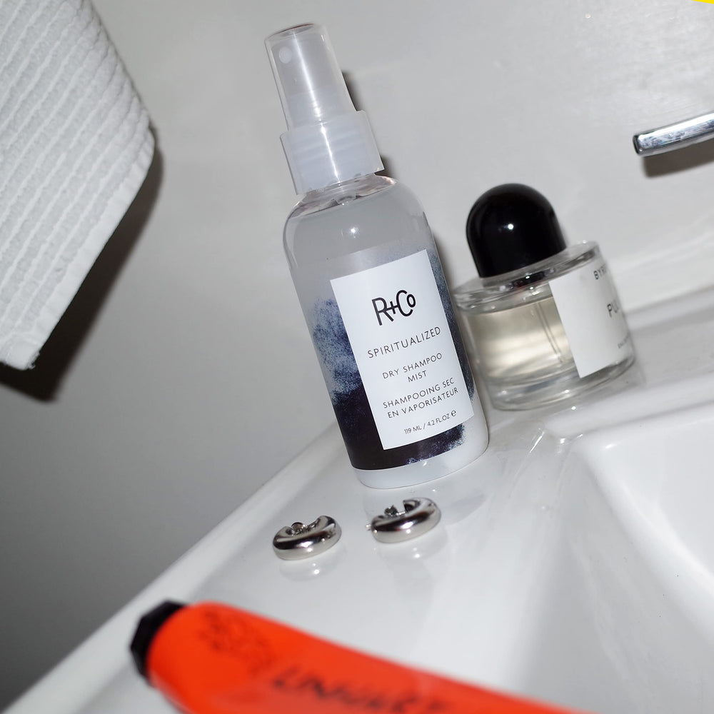 Hands applying R+Co Spiritualized Dry Shampoo Mist to roots to activate cleansing.